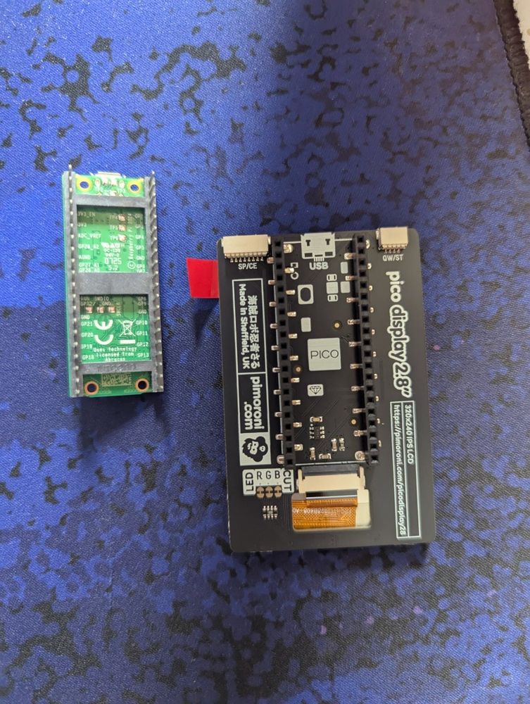 The back of a Pimoroni Pico Display Pack 2.8", its sockets for Pico headers facing upward; the camera casts down upon it and a green Raspberry Pi Pico 2 W, but looks on at a slight angle.

The Raspberry Pi Pico 2 W board beside it has its headers facing similarly upwards, but rests tilted, its headers leaning towards the Display Pack sat around a centimetre away.

Visible is the 'USB' indicator printed to the bottom of the Pico Display Pack, which informs the user as to how to orient their Pico while attaching it to the bottom, so the GPIO pins are connected properly and not in a flipped order.

Both sit on a blue-ish section of an LTT desk pad, roughly towards the bottom centre of its design.