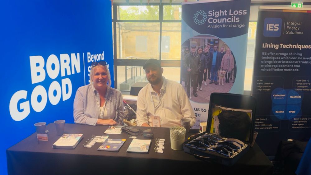 Photo Description:
Two people are seated behind a table at an indoor event. On the left is a smiling woman with blonde hair wearing a striped shirt. On the right is a man with a beard wearing a white shirt and black cap. Behind them are three large banners. One bright blue banner says “BORN GOOD | Beyond Sight Loss.” Another banner is for “Sight Loss Councils” with a photo of a diverse group of blind and partially sighted people, including a guide dog. The third banner promotes IES (Integral Energy Solutions). On the table are leaflets, keyrings, cups, and a small suitcase displaying simulation glasses used for sight loss awareness.

