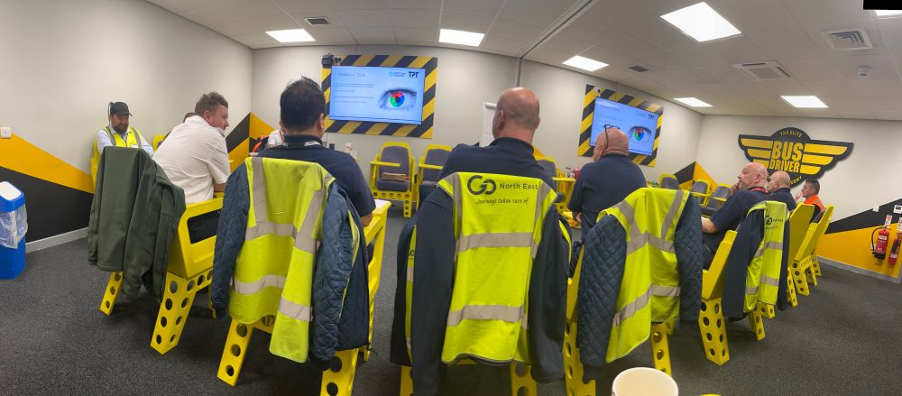 A group of bus drivers wearing high-vis jackets sit in a bright training room, facing a presentation screen. The walls are decorated in yellow and black with “Bus Driver Academy” branding.