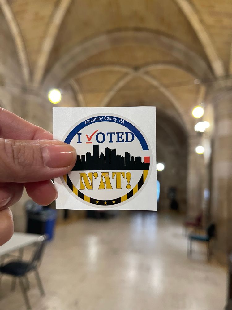 My “I voted ‘n ‘at” sticker held in my thumb and index finger. The arches of the courthouse lobby is in the background. 