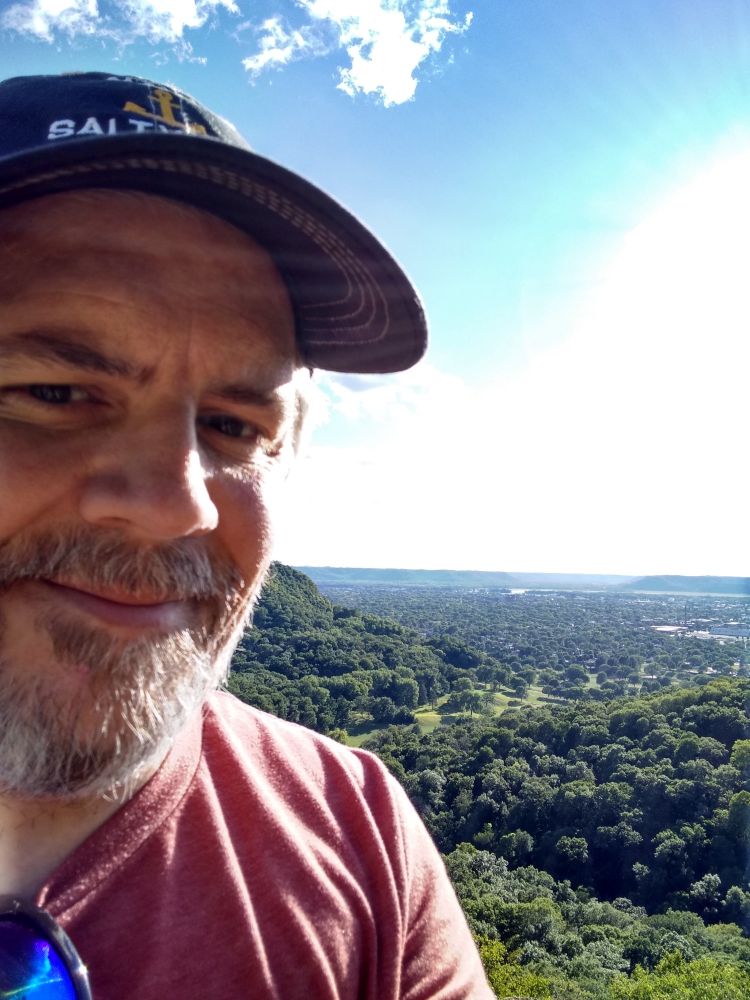 White guy in a ball cap with a grey beard. Over the left shoulder a view of the Mississippi valley from the top of a bluff. 