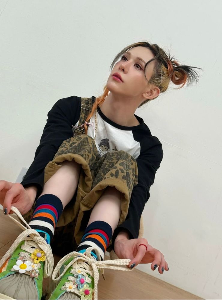 wumuti in leopard print overalls over a black and white longsleeved tee. she has colorfully striped socks and green shoes with flower charms on the laces. she has a cute little braid in her hair and she is looking up and to the side with a little puppy expression