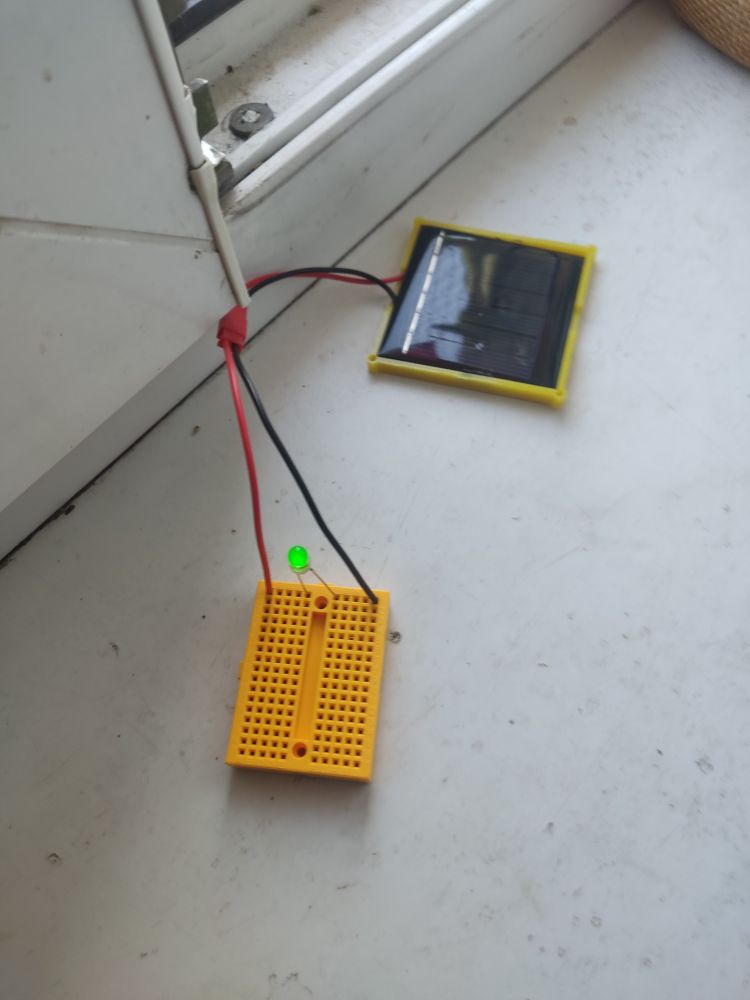 Układ elektroniczny na płytce konceptowej, składający się z panelu fotowoltaicznego i jednej diody świecącej, leży na parapecie warsztatu i świeci 