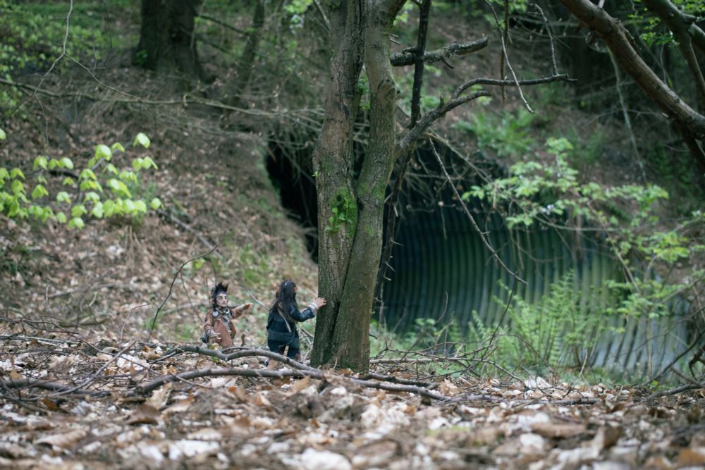 A photo of the Wee Folk looking towards a large culvert. The wee folk are two freestanding handmade puppets. They’re dressed in layered, homemade looking tweedy clothes, with lots of belts and gondoliers of useful items. They have pointed ears and large hands.