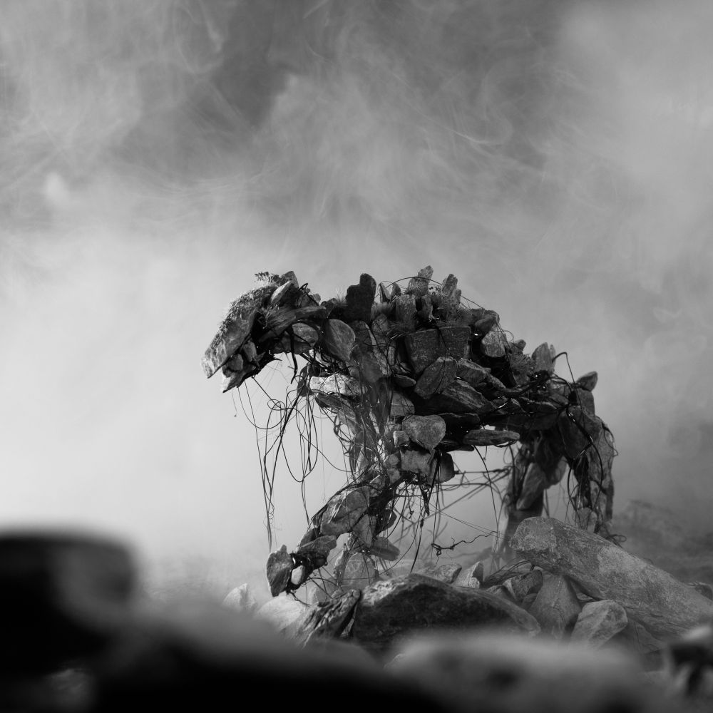 A monochrome image from the Pine Pitch Black edition of Maskwitches. A quadrupedal spirit made of stones, covered in black threads advancing through a rocky landscape. 