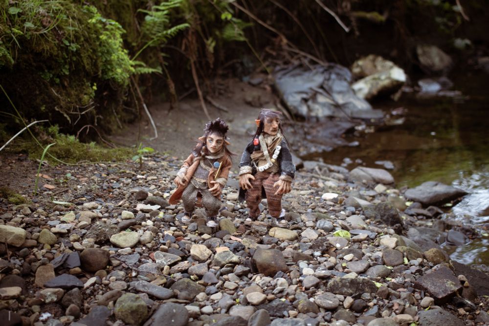 A photo of the Wee Folk making their way along the burn. The wee folk are two freestanding handmade puppets. They’re dressed in layered, homemade looking tweedy clothes, with lots of belts and gondoliers of useful items. They have pointed ears and large hands. They’re wearing little converse style shoes. The bank of the burn is a pebble beach. 