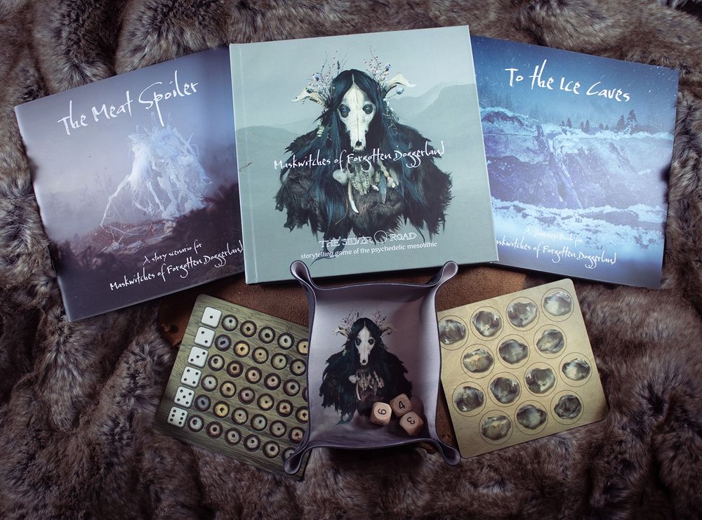 A photo of the Maskwitches range, showing the standalone core book, the Meatspoiler scenario, the To the Ice Caves supplement, the predestined event tokens, Knap tokens and a Maskwitches dice tray. 