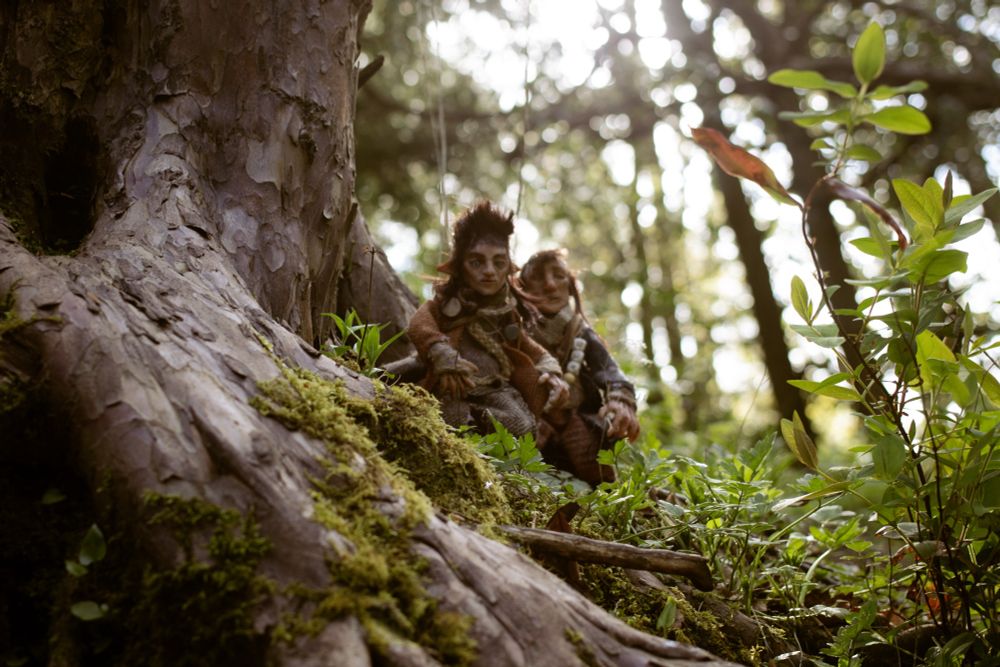 A photo of the Wee Folk making their way along the burn. The wee folk are two freestanding handmade puppets. They’re dressed in layered, homemade looking tweedy clothes, with lots of belts and gondoliers of useful items. In this image the wee folk are descending a slope at the foot of a big tree making their way through the roots. 