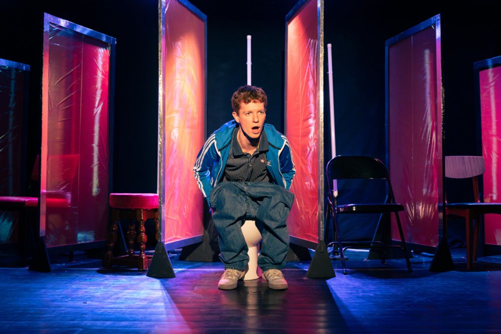 A man in blue jacket sitting on a toilet with his trousers pulled down. He looks surprised with his mouth open. 