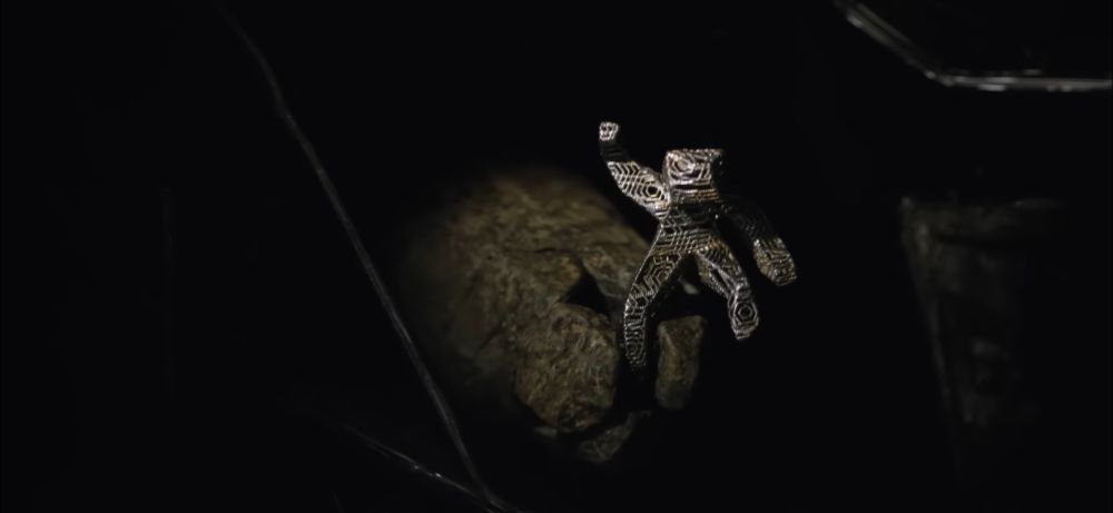 A little metal stick man sculpture of an astronaut is grasped by three rocky pincer fingers emerging from the darkness.
