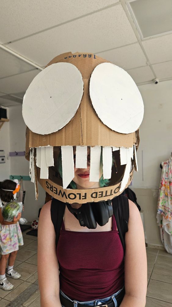 a child wears a cardboard mascot like mask about 4 times the size of their head. The mask is round has a large mouth, which the wearer can see through, with square teeth of varying lengths. The eyes on the mask are huge, circular, and white, with tiny pin pick pupils. 