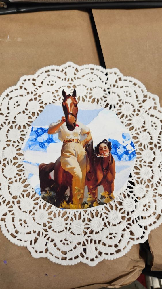 A collage of a woman standing beside her horse, but their heads have been swapped. The figures are laid on a sky of textured paper, one piece is bubble painted, and other are torn blue or white pieces. The piece is mounted on the centre of a doily. 