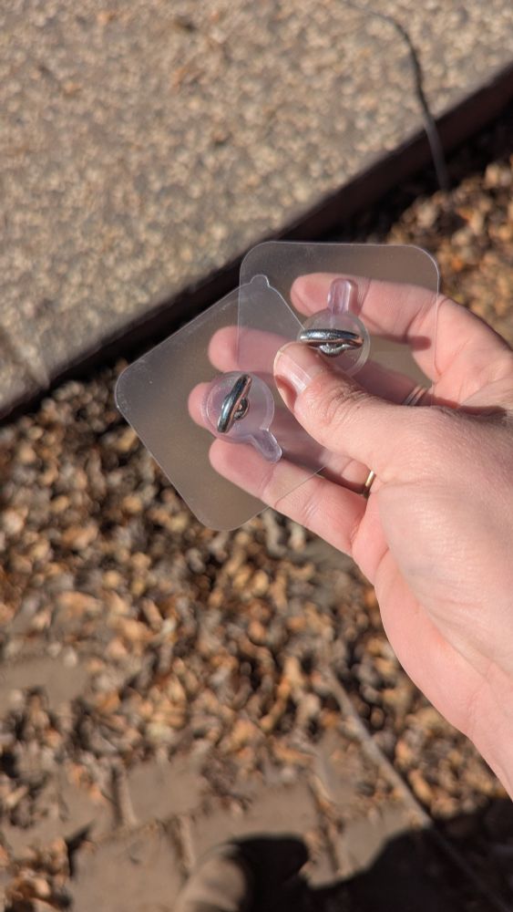 A white woman's hand holding two clear plastic adhesive pads with a small silver metal eye bolt mounted in the center of each one.