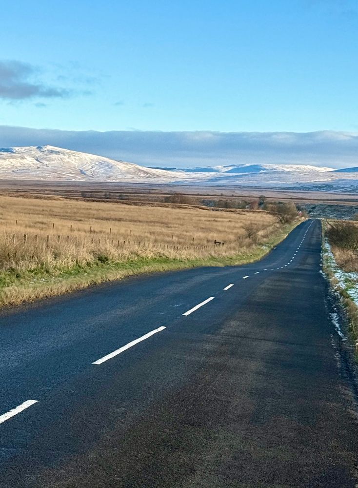 A tarmac road undulates through a grassy moorland to snow covered hills and blue sky beyond. Original photograph by me. Ayrshire, Scotland. 