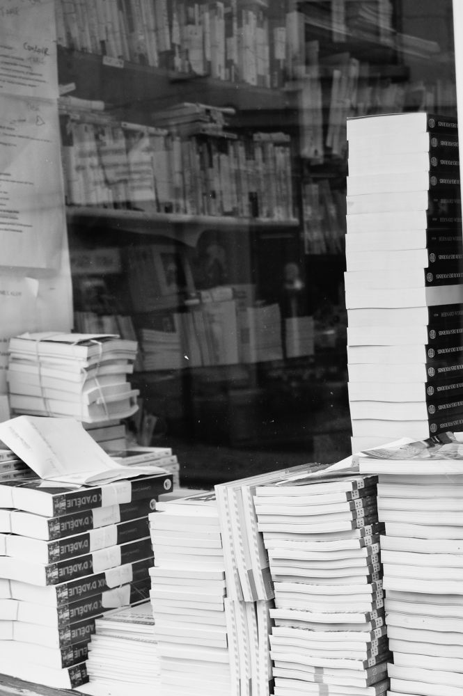 Photo en noir et blanc d’une devanture de librairie montrant des piles de livres.