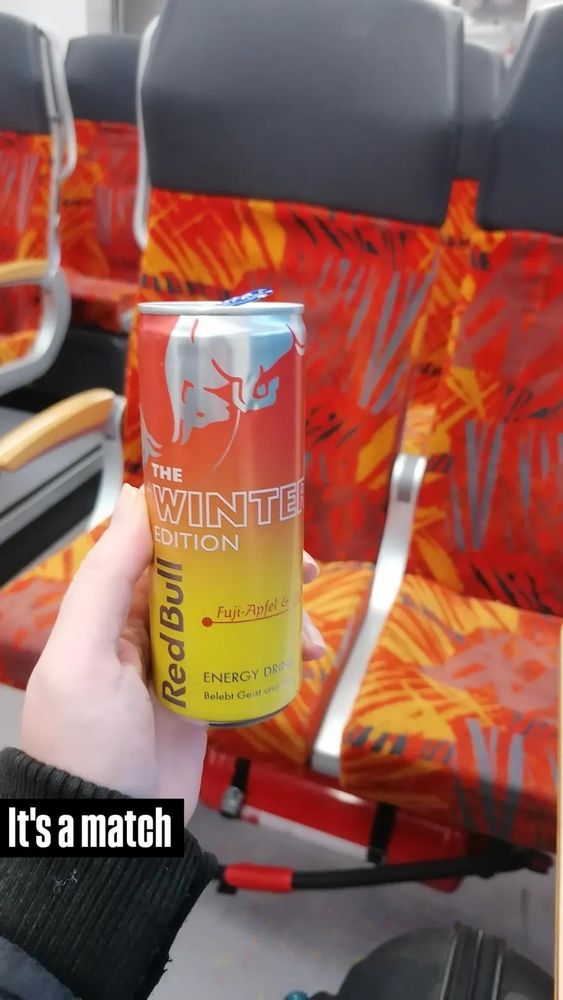 Im Vordergrund wird eine Dose Redbull in die Kamera gehalten welche im Farbverlauf von Rotorange zu Gelb wechselt (gelb unten) im Hintergrund sind Zugsitze zu sehen, deren Bezug praktisch die gleichen Farben aufweist.
