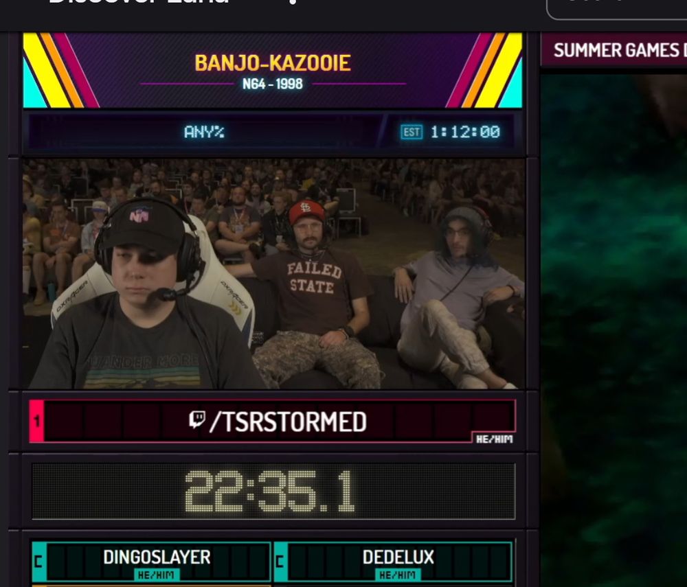 a screen shot from a live stream of “summer games don quick” a man with a headset sits in the foreground, two people are sitting on a coach behind him watching. one person is wearing Cola merch that says “Failed State”