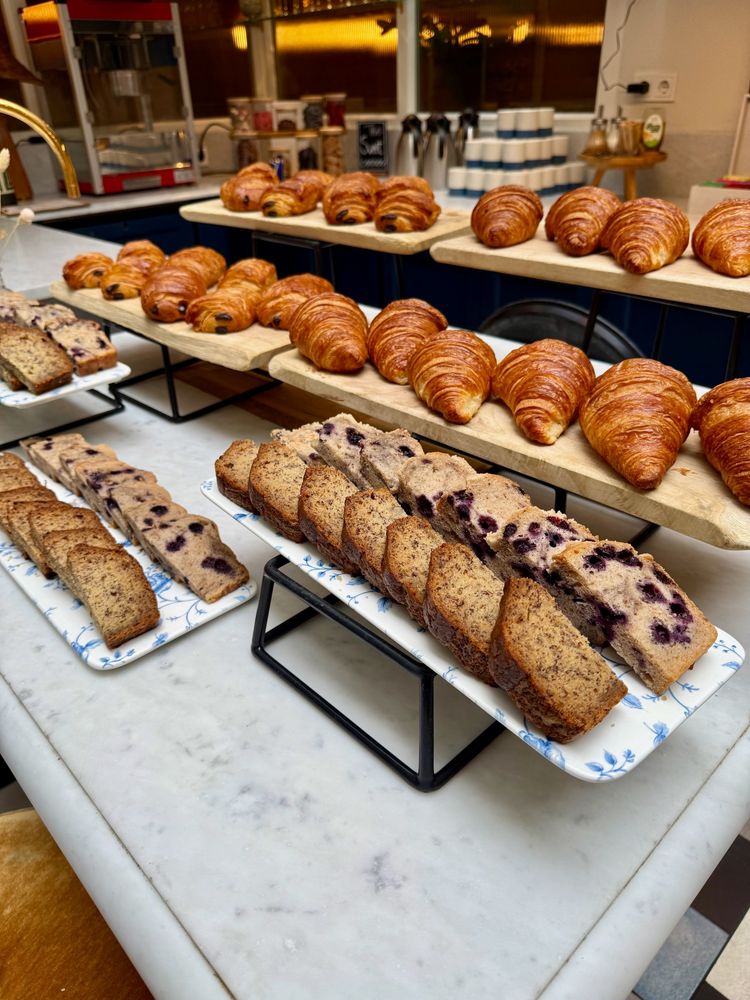 A breakfast buffet featuring rows of pastries and sliced cakes. The front trays display banana bread and blueberry loaf slices on floral-patterned plates. Behind them, wooden boards hold neatly arranged croissants and pain au chocolat.
