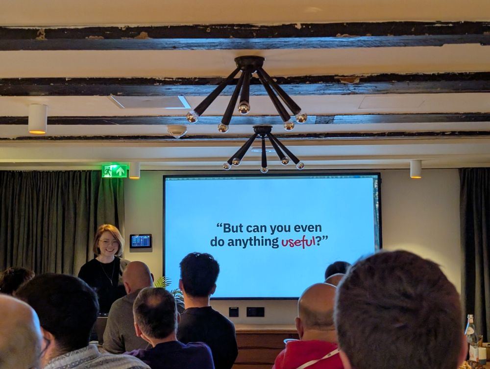 Julia stands at the front of a cozy, wood-beamed room giving a talk to an attentive audience. Behind her, a presentation slide reads: ‘But can you even do anything useful?’