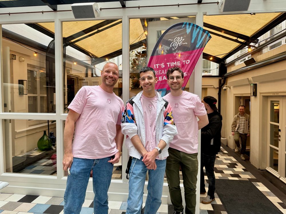 Nils, Nesim, and Jan (from left to right) stand smiling in front of a CSS Café banner. All three are wearing light pink shirts with a small owl graphic. The setting is a covered courtyard with checkered floor tiles and glass walls. A few other people are visible in the background.