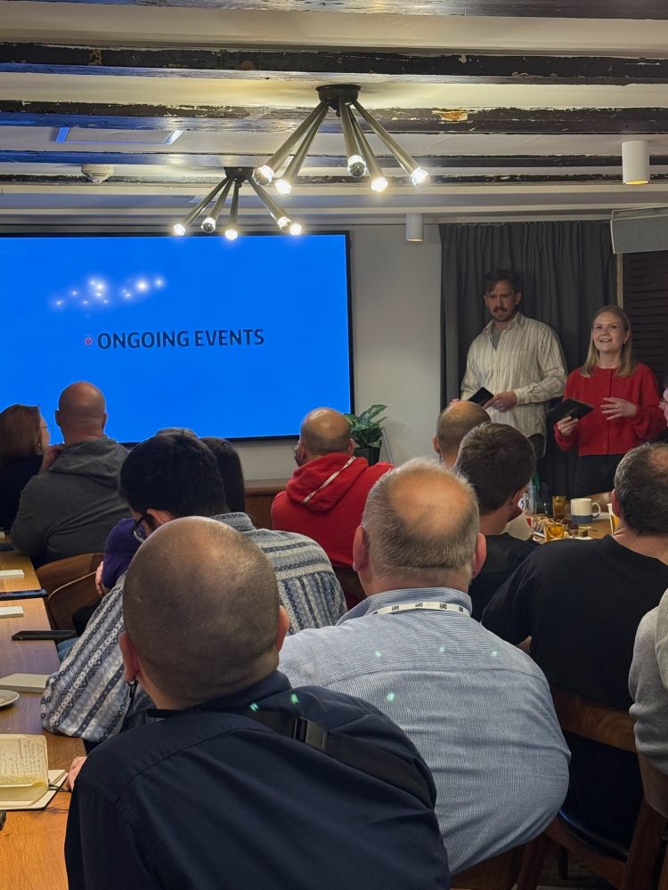 Johannes and Vilde are presenting in front of an audience at an indoor event. A large screen behind them displays the slide title “Ongoing Events.” The room is filled with seated attendees, viewed from the back, and has modern ceiling lights and wooden beams.