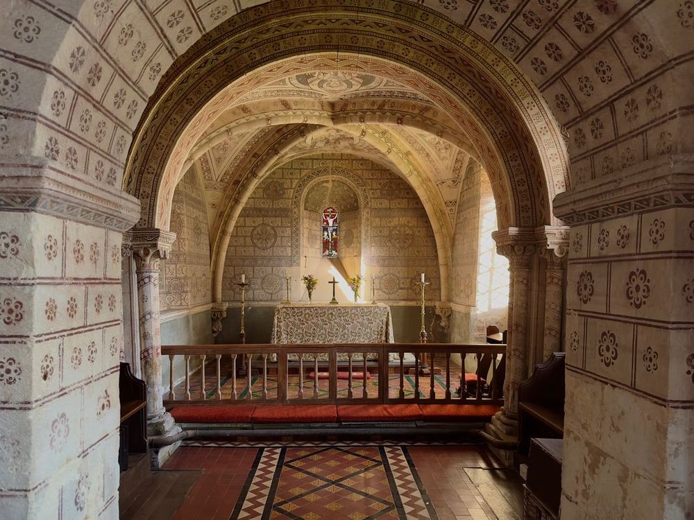 Interior of St George’s Church, Hampnett, Gloucestershire: richly painted Norman chancel with geometric wall patterns, arched ceiling, tiled floor, and an altar lit by stained glass.