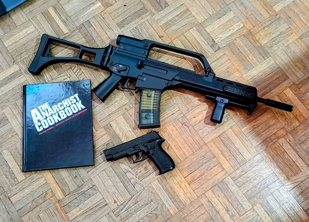 Un livre "The Anarchist Cookbook" et une réplique airsoft de G36KE et une de Sig-Sauer P226 posés sur le parquet de mon salon. 