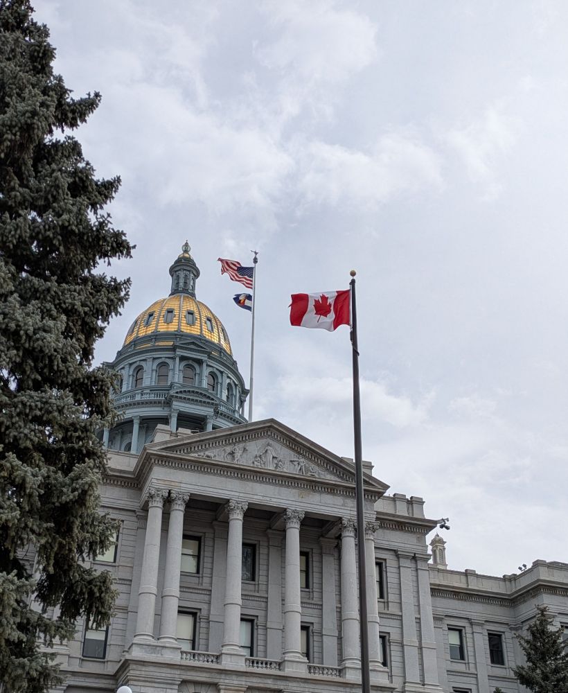 The Canadian, U.S., and Colorado flags fly in front of the hills dome of the Colorado State Capitol building.