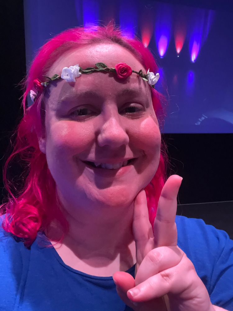 A person with pink hair and a flower crown smiling at the camera and holding up a peace sign with their left hand. They’re wearing a blue shirt.