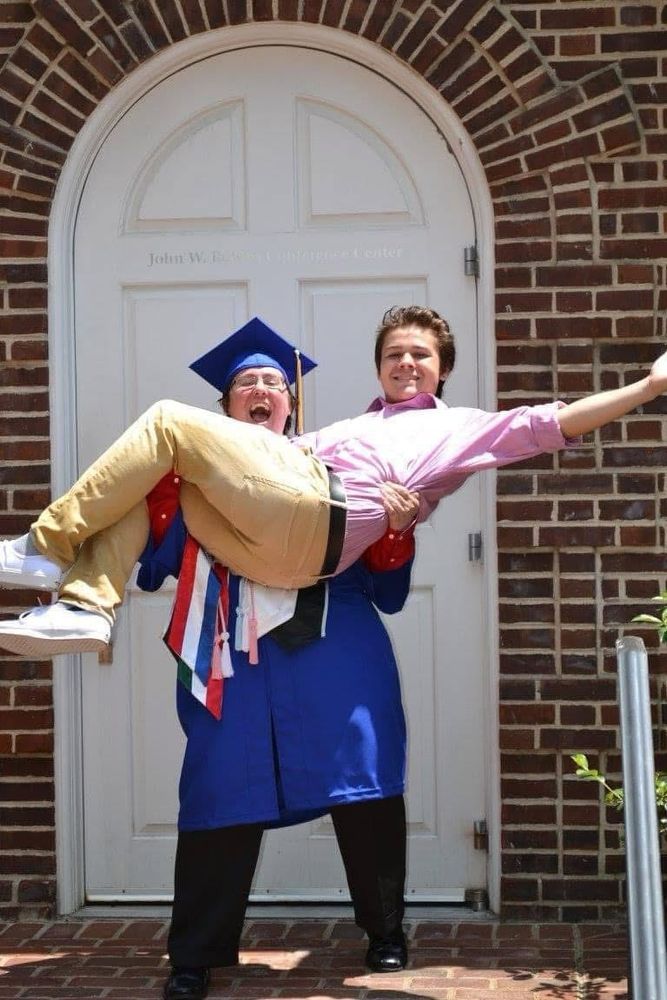 A person in a graduation lifting up a teenage boy in a pink shirt and khakis