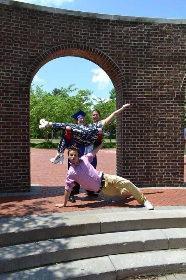 A person in a graduation gown lifting a girl who is wearing a patterned jumpsuit and chunky white heels and a teenage boy is posing in front of them wearing a pink shirt and khakis