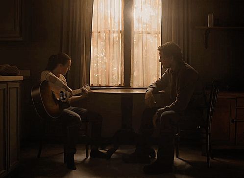 a woman is playing a guitar while a man sits at a table in front of a window