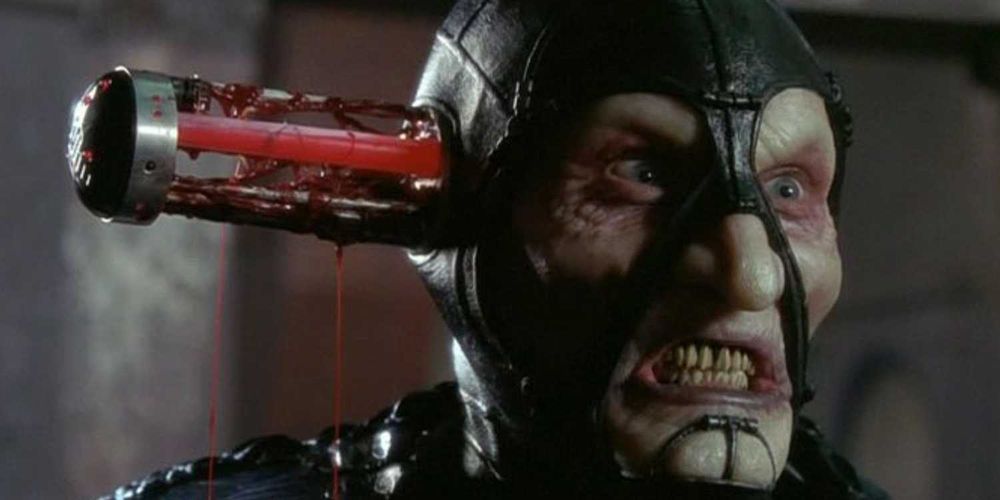 Screen capture of Scorpius (Wayne Pygram) from Farscape, a man with translucent semi-reptilian skin over skeletal features, wearing a leather cowl and alligator-skin armor. The ear cap of his temperature control suit has popped out, revealing the spent cooling rod from inside his brain. The housing is oozing red goo.