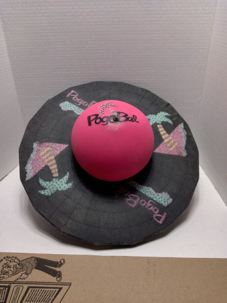 Image of a vintage Pogo Bal, a bright pink ball reading Pogo Bal in black lettering over a green and yellow palm tree mounted on a black plastic platform with an 80s triangle and palm tree motif (green and yellow palm tree, pink triangle) printed on it. Another pink ball (unseen) is mounted on the opposite side of the platform for bouncing. 