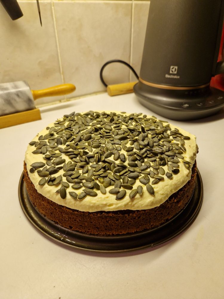 A pumpkin cake on a kitchen counter, cream cheese frosting with a cover of dry roasted pumpkin seeds. In the background, an electric water kettle with temperature settings and a keep warm function that I would love would it not beep all the time.