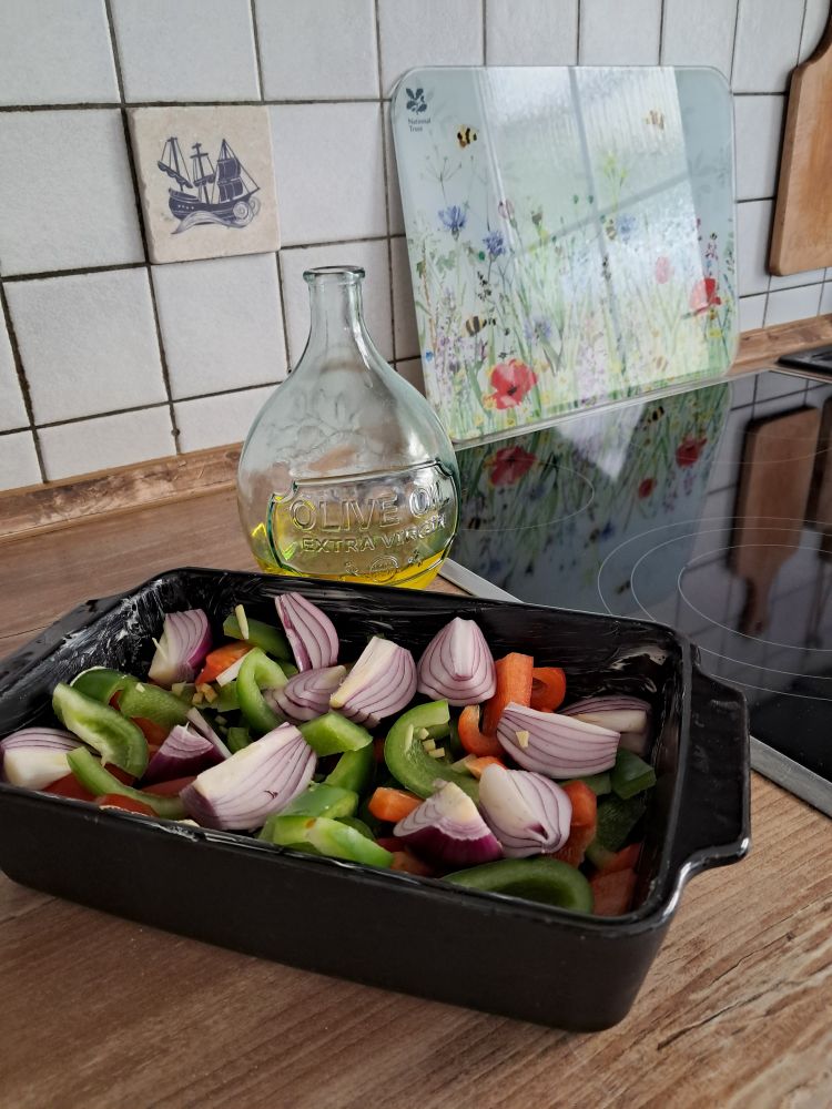 Neben dem Herd steht eine schwarze Auflaufform mit Paprika- und roten Zwiebelstücken, dahinter eine Karaffe mit Olivenöl.