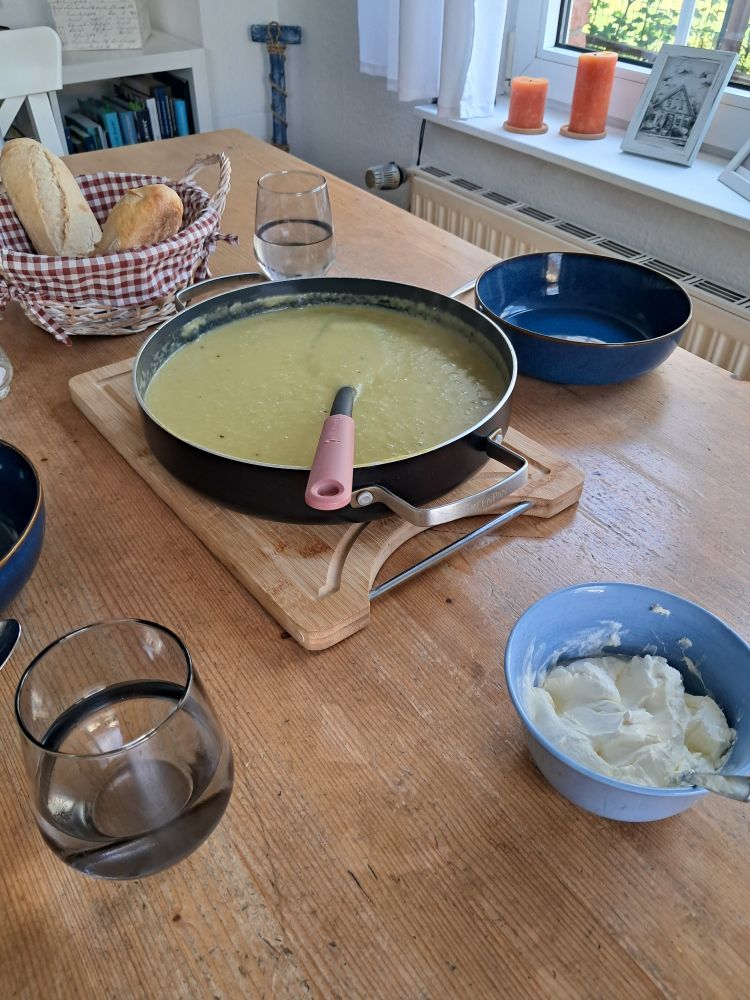 Ein Holztisch mit einem Topf grünlicher Suppe, zwei Tellern, Brot und einem Schälchen mit Schmand.