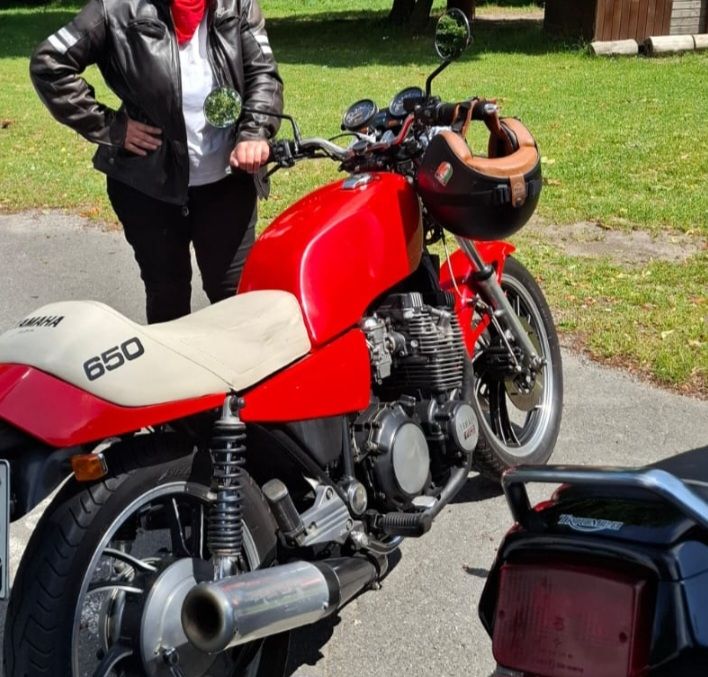 Ein rotes Motorrad mit weisser Sitzbank, dahinter steht die Hüterin mit einer Hand am Lenker. Gesicht und Nummernschild sind nicht zu erkennen