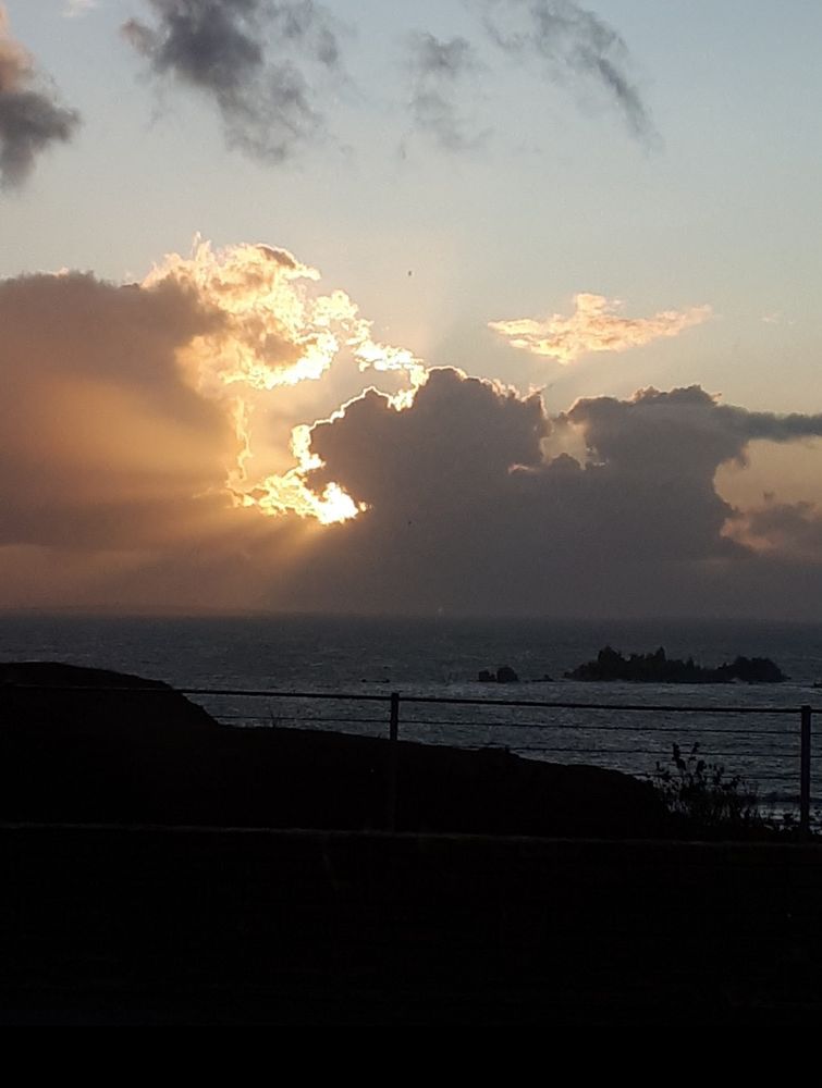 Dämmerung, hinter einem einfachen Zaun liegt das Meer, ein paar Felsen ragen daraus hervor. Am Himmel verdecken dunkle Wolken die Sonne fast ganz,etwas Licht und einige Sonnenstrahlen sind zu erkennen. 