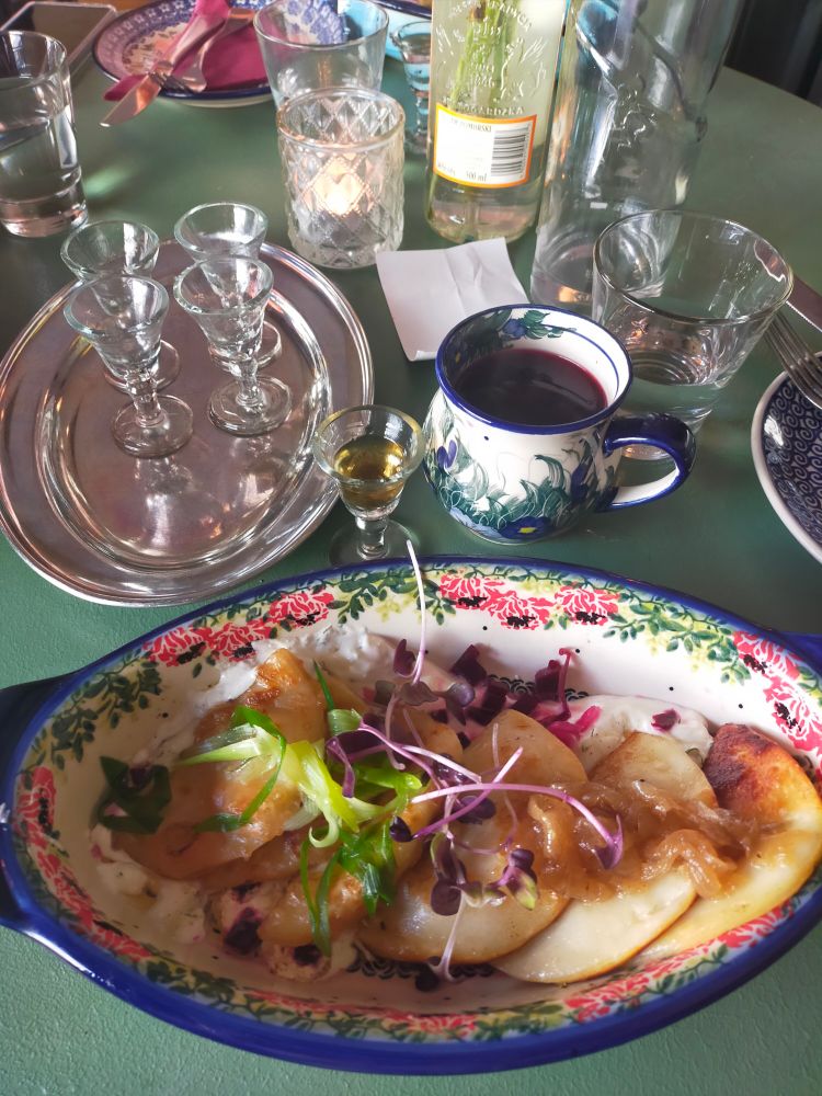 A dinner setting. An oval bowl of potato and cheese pierogi, served with sour cream, Caramelised onion and garnished with flowers. Some empty vodka shots on a small tray. A cup of borscht. Bottles of water and glasses in the background. A cheerful display.