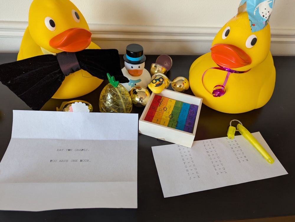 A task reads EAT THE GRAPES. YOU HAVE ONE HOUR. Around it sit a rainbow puzzle, some golden eggs, a plastic pineapple, and four rubber ducks of varying sizes, as well as a cipher and invisible ink pen and light.