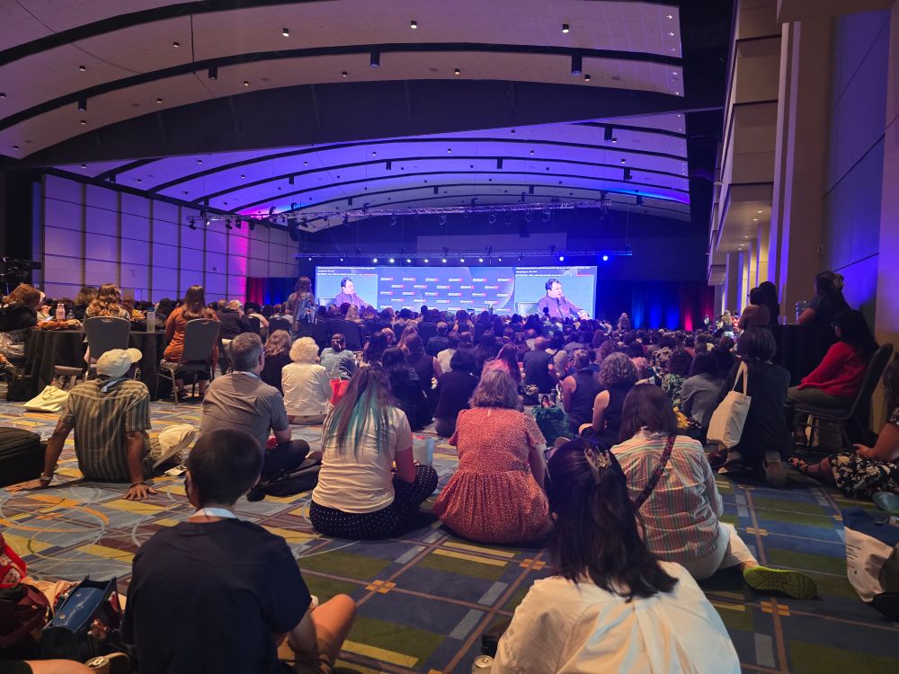 A crowded ballroom with people watching a presenter.