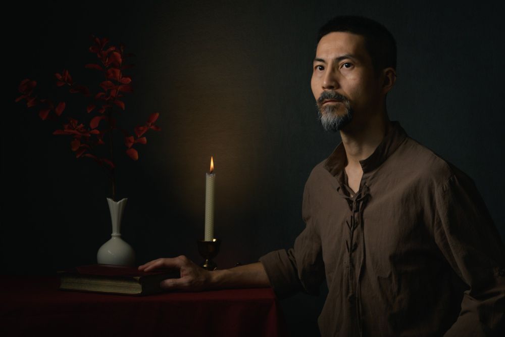 This is a portrait of a middle-aged Japanese man wearing a brown Renaissance-style shirt on the right side of the photo with his body turned to the left. He is sitting at a table draped with red cloth and looking toward the left. His right hand is placed on the table touching an old book. In the background, there is a lit candle that lights the wall. To the left of the candle is a white vase with a small tree branch with red leaves.