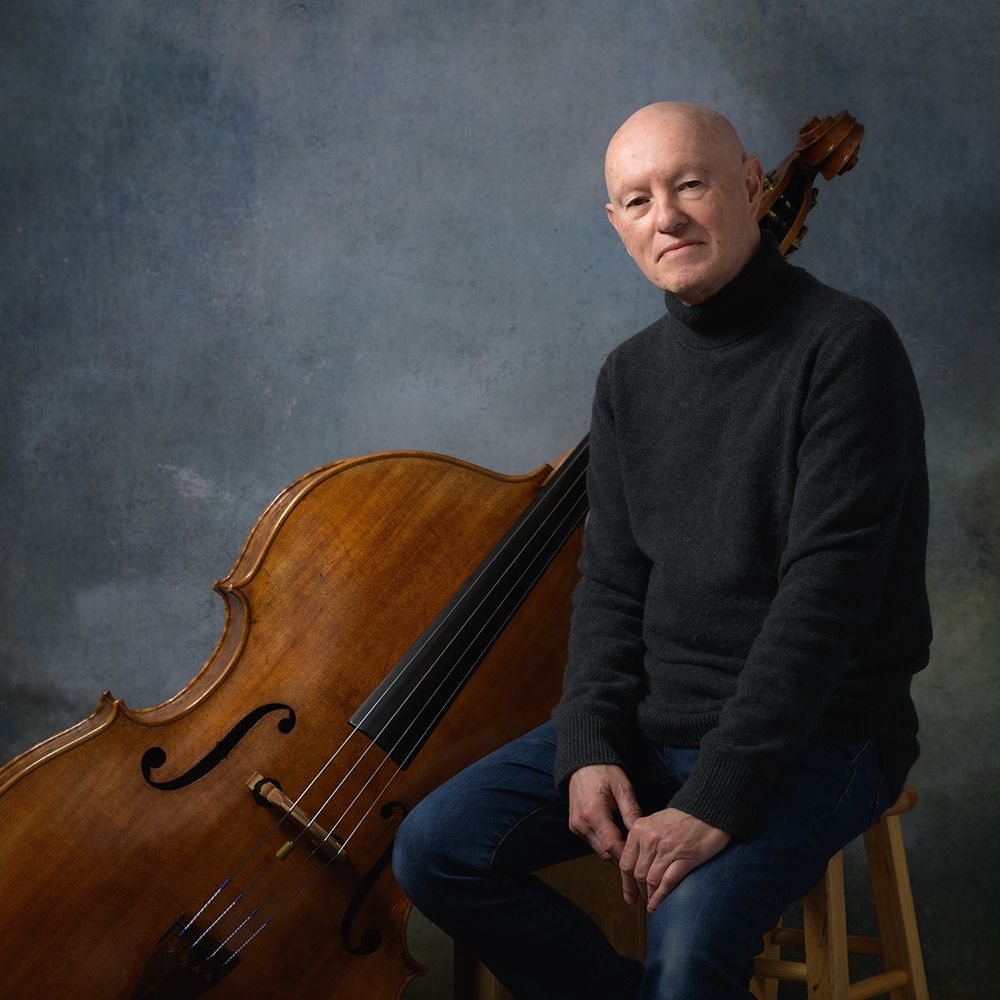 This is a portrait of Mark Bernat with his double bass. Mark is a caucasian man wearing a dark gray turtle neck sweater and a pair of blue jeans, sitting on a stool and looking at the camera. His bass is placed behind him, leaning against a wooden box. Background is designed to look like a classical painting of blue-gray sky. 