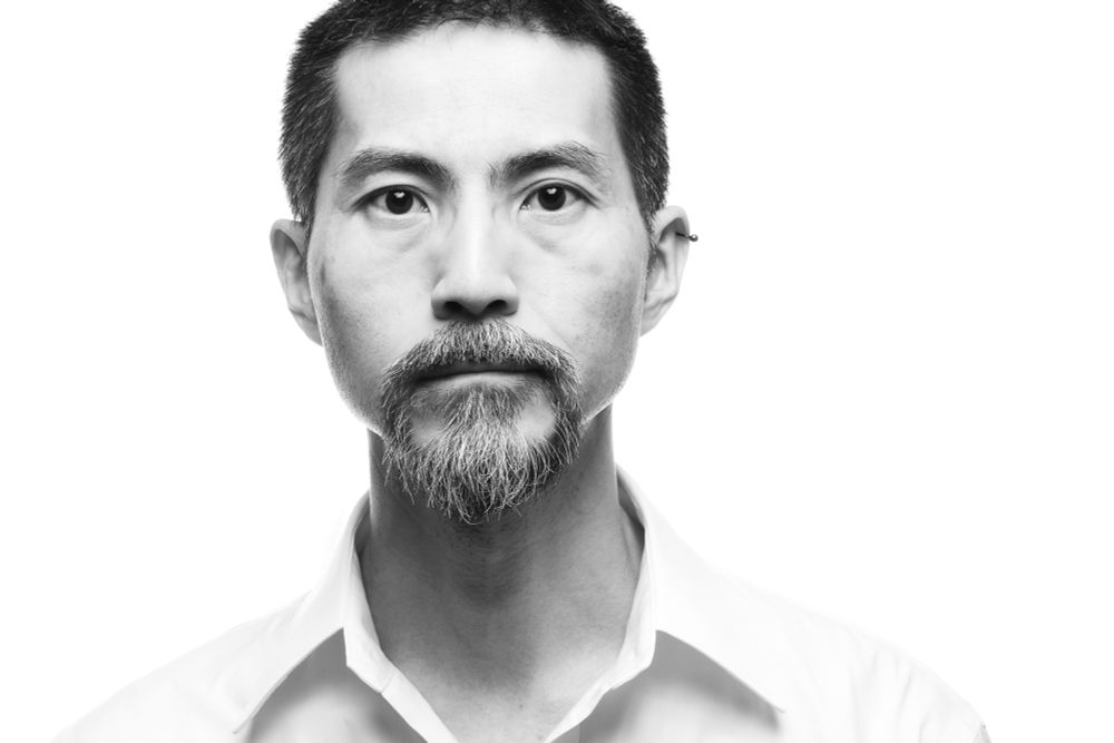 This is a high-key portrait of a middle-aged Japanese man wearing a white shirt. He is looking straight at the camera. The background is white.