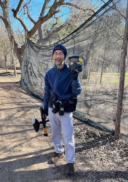 A photo of a middle aged Japanese man holding a paintball mask with a yellow paint splatter.