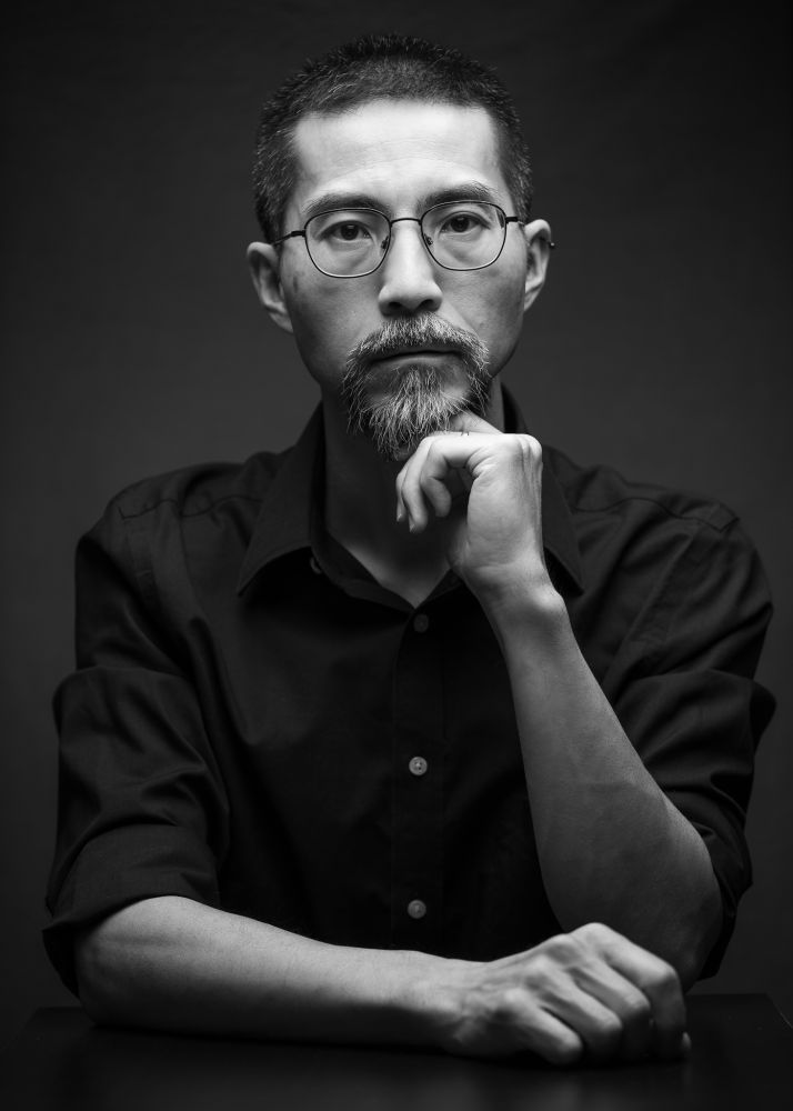 This is a black-and-white portrait of a middle-aged Japanese man wearing a dark shirt. He is wearing glasses and looking straight at the camera. He is sitting at a table with his right arm out in front and the left hand under his chin. The background is dark gray with a little bit of lighting.