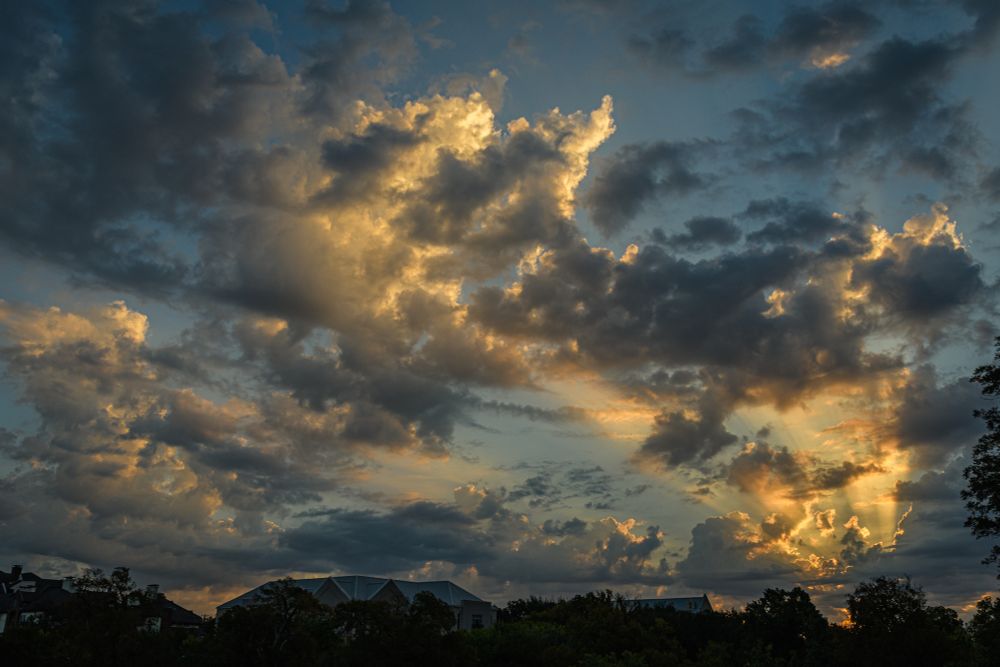 This is a photo of a dramatic sky during sunrise as seen over a residential neighborhood in Austin, Texas. As the sun rises from the lower right of the image, beams of sunlight stream through and illuminate fast-moving clouds.