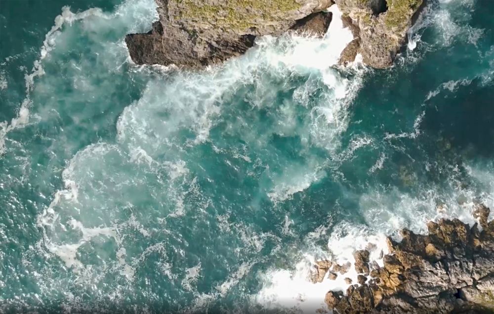 A dramatic aerial view of turquoise waves crashing against rugged cliffs, swirling with energy and white foam. A wild and powerful seascape, alive with motion, this is the marine world we must protect.