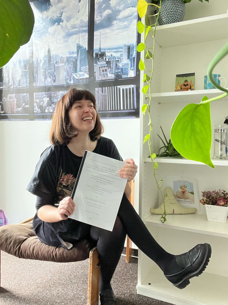 Julia sits in a chair in her office and holds her printed thesis. She is laughing and not looking into the camera. Julia looks relieved. Green leaves of plants surround the photo and dangle from a cupboard next to her. Behind her is a wall poster with an aerial view of New York City.